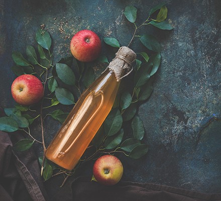 Rustic looking bottle of apple cider vinegar lying next to some apples and apple leaves on a dark, mottled surface