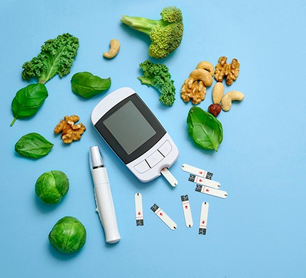 A blood glucose monitor surrounded by healthy green foods and nuts, on a blue background