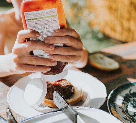 Ketchup bottle - with a label of many different ingredients clearly visible - being squeezed over a burger