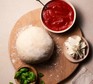 Pizza dough on a board next to a bowl of tomato sauce and a ramekin of basil